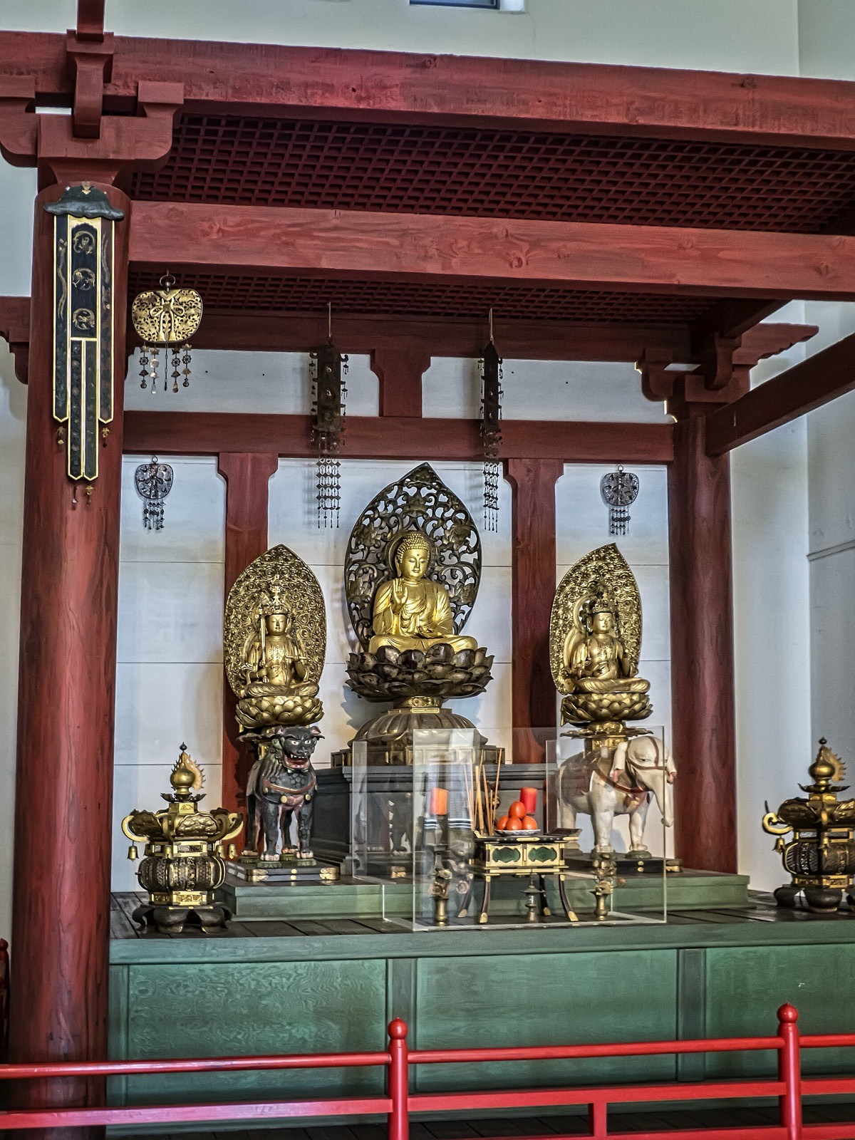 El santuario interior de un templo budista Shingon: tres figuras búdicas doradas sobre pedestales de loto bajo columnas bermellón, con un par de velas de pilar naranjas, varillas de incienso y frutas de ofrenda en la mesa de delante.